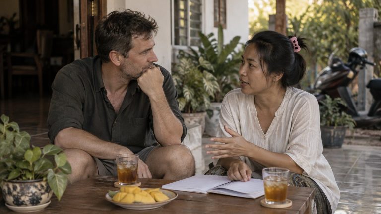 A Western man and a Thai woman talking over a notebook at a table on the porch of a Thai family house